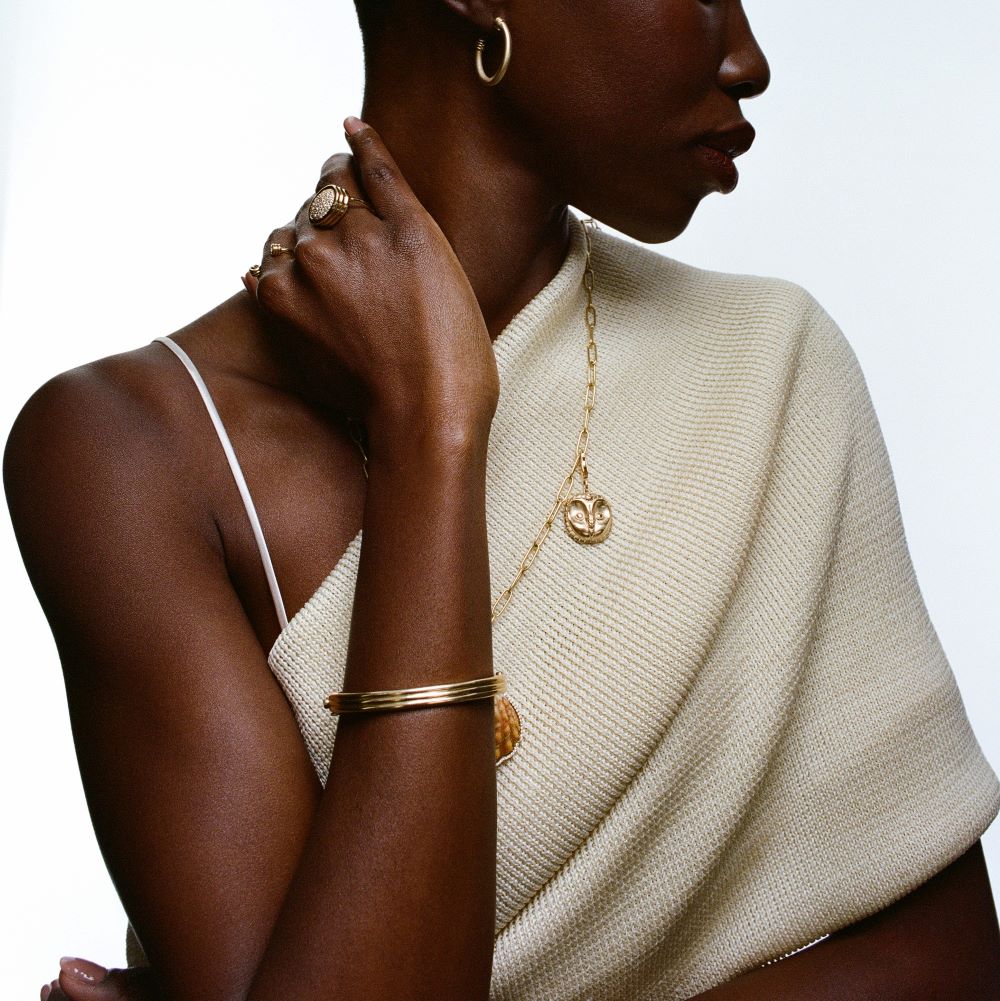 A black woman wearing a white sweater and gold jewelry, including a Futura Peruvian Moche Owl Charm.
