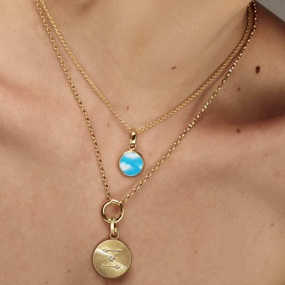 A woman is wearing an Anna Maccieri Rossi hand-painted gold necklace with a blue Art Dreamy Charm pendant inlaid with Australian Mother of Pearl.